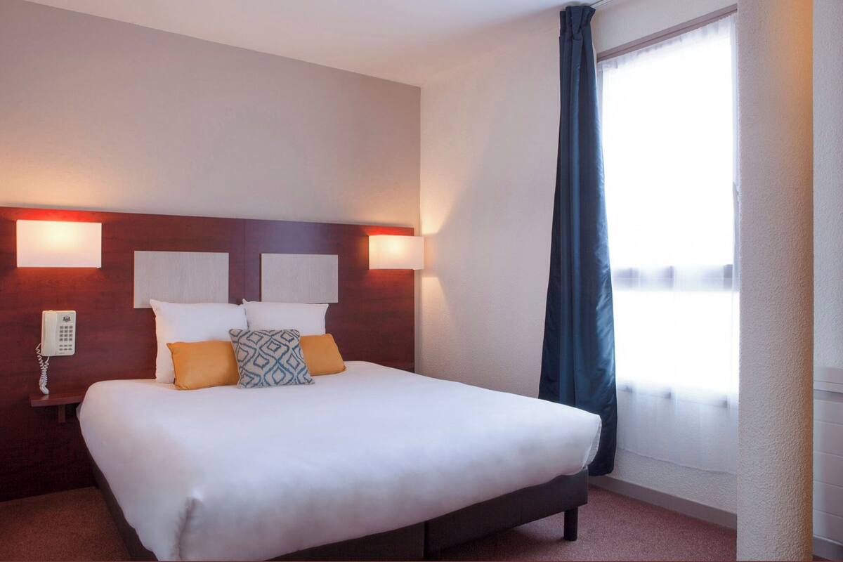 Ein Hotelzimmer mit einem großen Bett, zwei Nachttischlampen, einem Telefon und einem Fenster mit Vorhang.