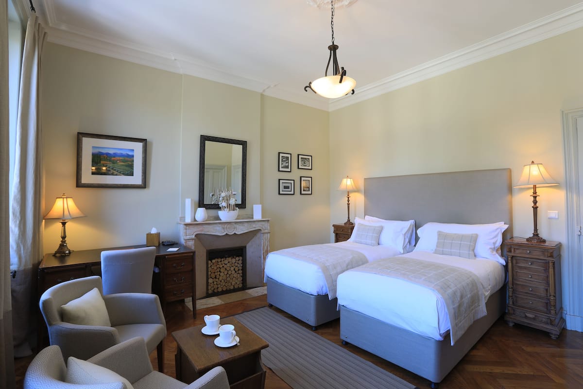 Una habitación de hotel con dos camas, un escritorio con lámpara, una silla, chimenea y cuadros en la pared.