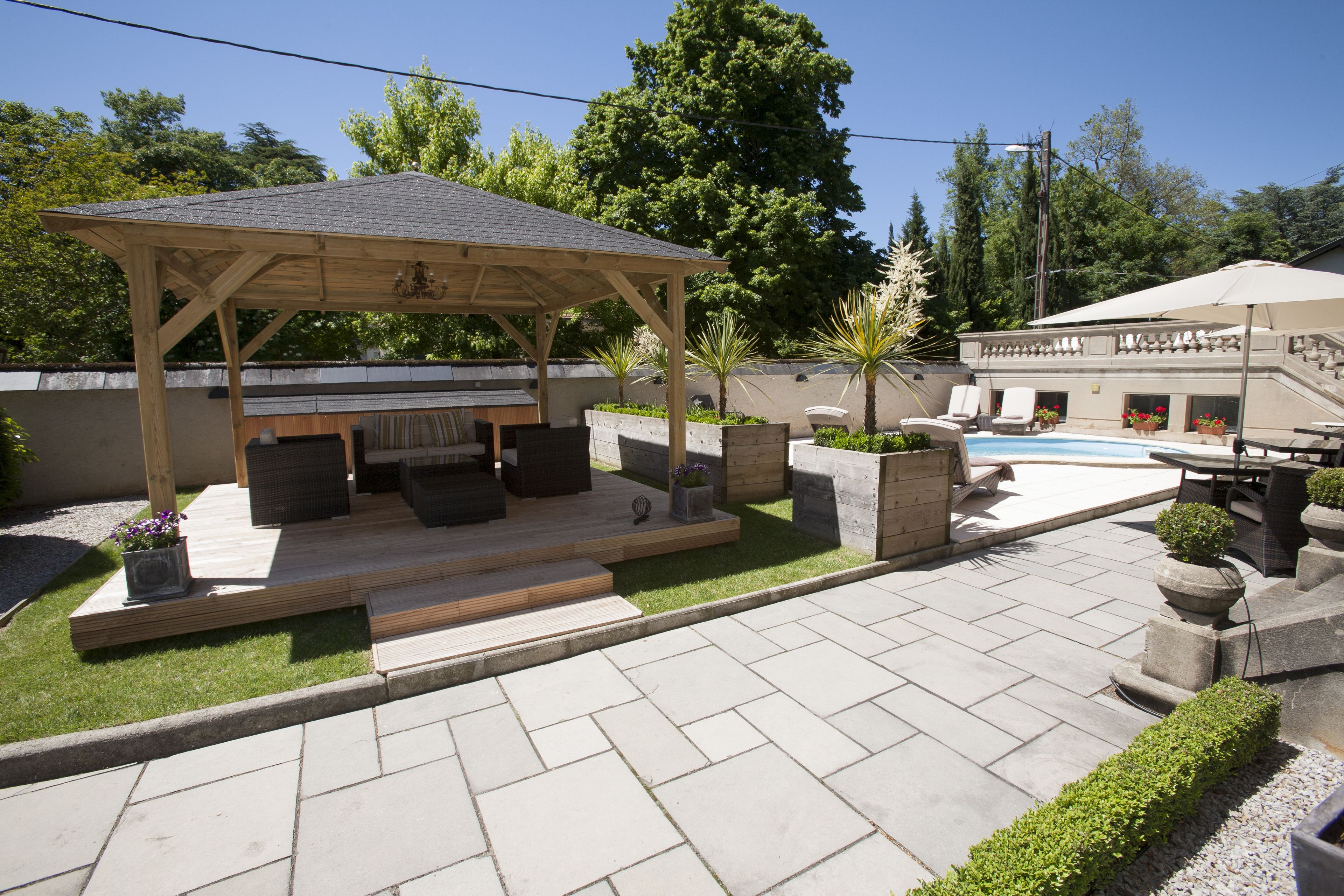 A covered outdoor seating area with a grill, surrounded by planters and a pool.