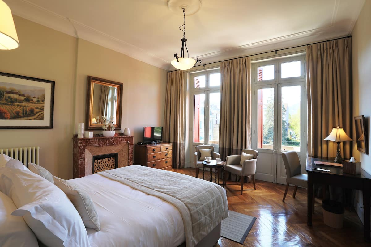 Habitación de hotel con una cama grande, chimenea, escritorio con lámpara y vistas al exterior a través de las ventanas.