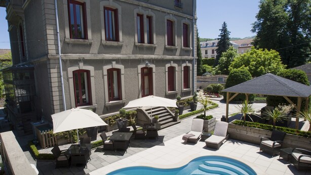 Un área de piscina con sillones de descanso y sombrillas, un edificio de piedra con contraventanas rojas y un cielo azul despejado.