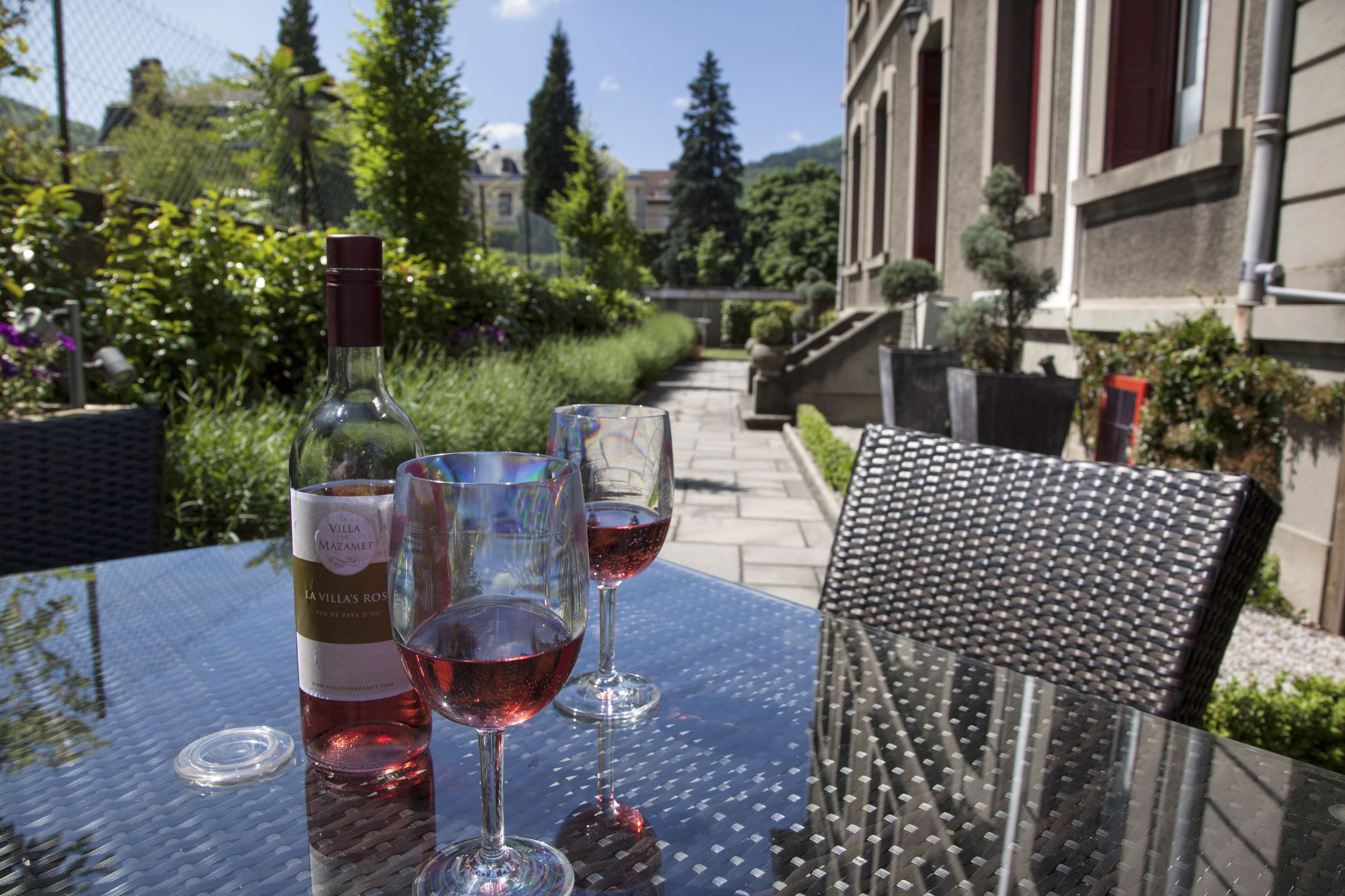 Una mesa con una botella y copas de vino tinto, colocada al aire libre con vistas a un jardín y edificios al fondo.