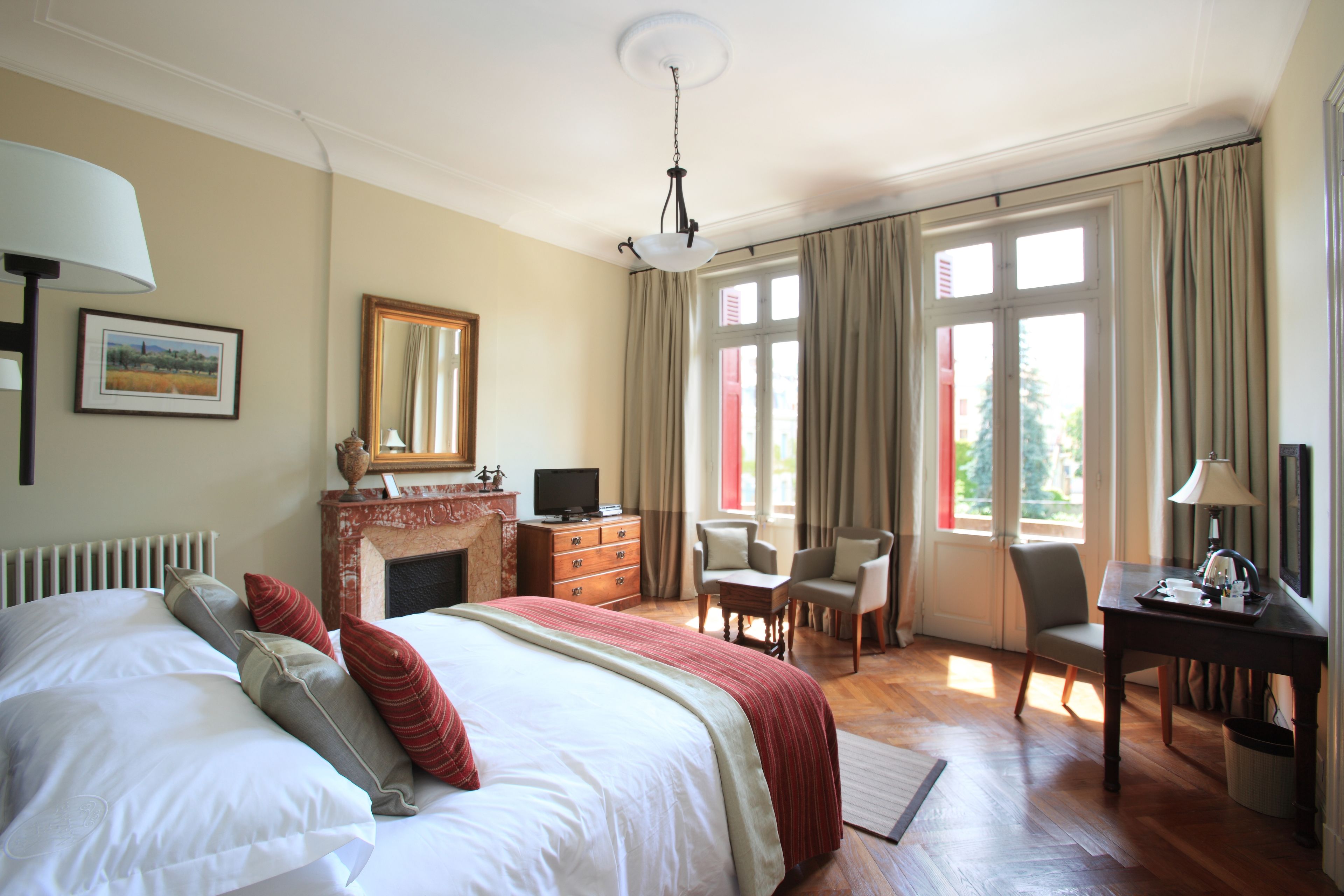 A hotel room with a large bed, a fireplace, a desk with a lamp, and a view of the outdoors through the windows.