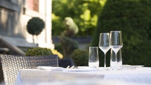 Mesa al aire libre con cristalería transparente y mantel blanco, rodeada de follaje y un edificio al fondo.