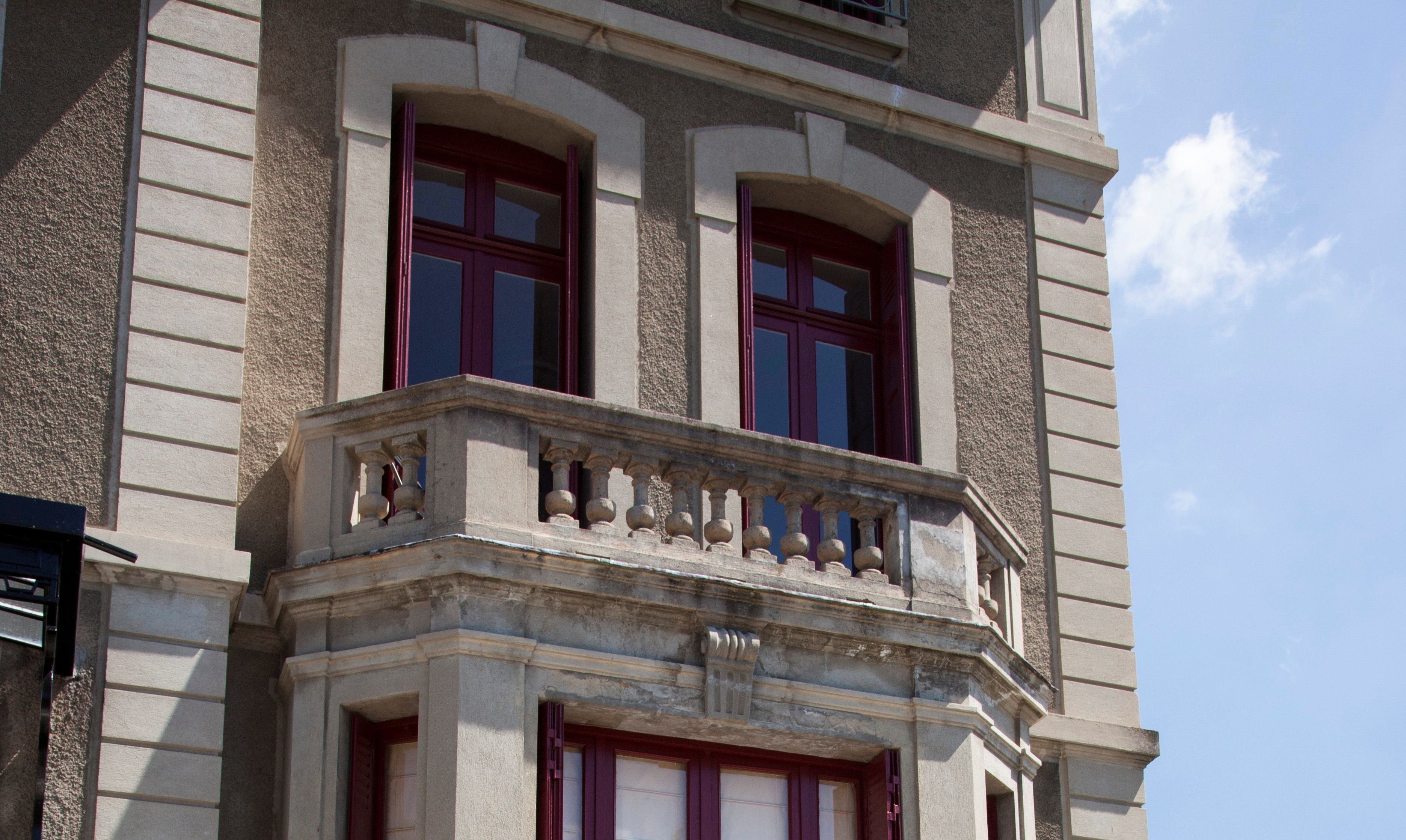 Un edificio con balcón que presenta balaustrada de piedra y dos ventanas con marcos rojos.