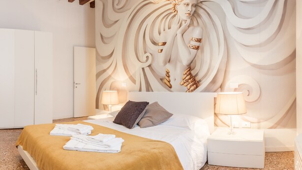 Une chambre d’hôtel comprenant un lit, deux lampes de chevet, une tête de lit ornée d’une fresque décorative, une porte et une table de chevet.