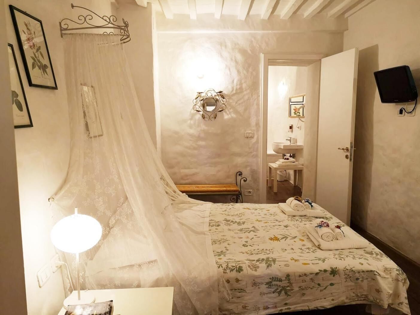 Un dormitorio con cama a dosel, una lámpara de noche, un espejo, un televisor y un baño visible a través de una puerta abierta.
