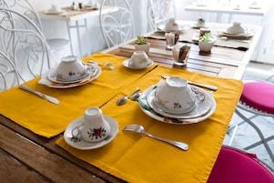 Una mesa de comedor servida con tazas y platillos blancos, cubiertos y un mantel amarillo.