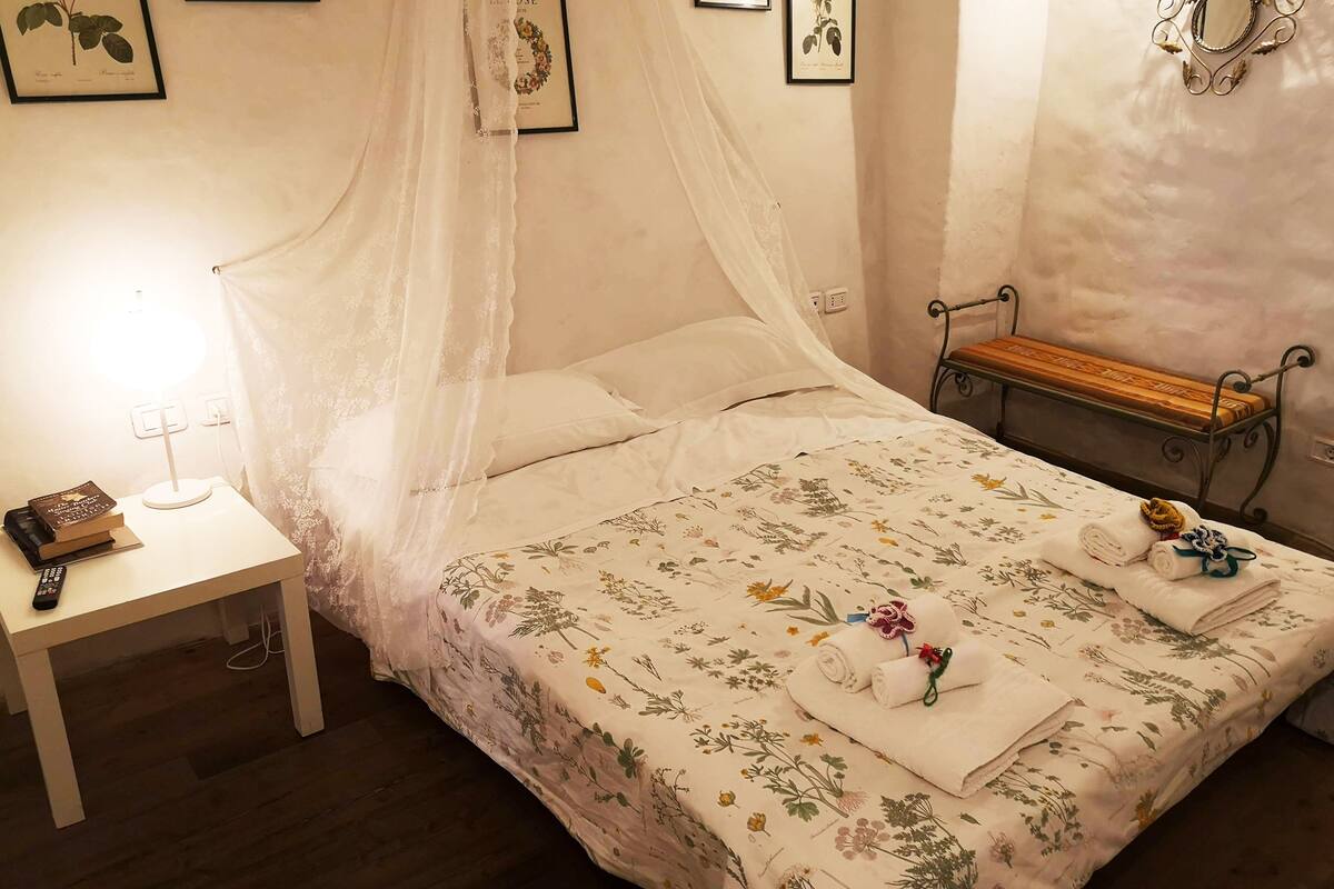 Una cama bien hecha con colcha estampada, una mesita de noche con lámpara, y un banco apoyado en la pared.