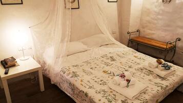 Cama bem arrumada com colcha florida, mesa de cabeceira com abajur e um banco encostado na parede.