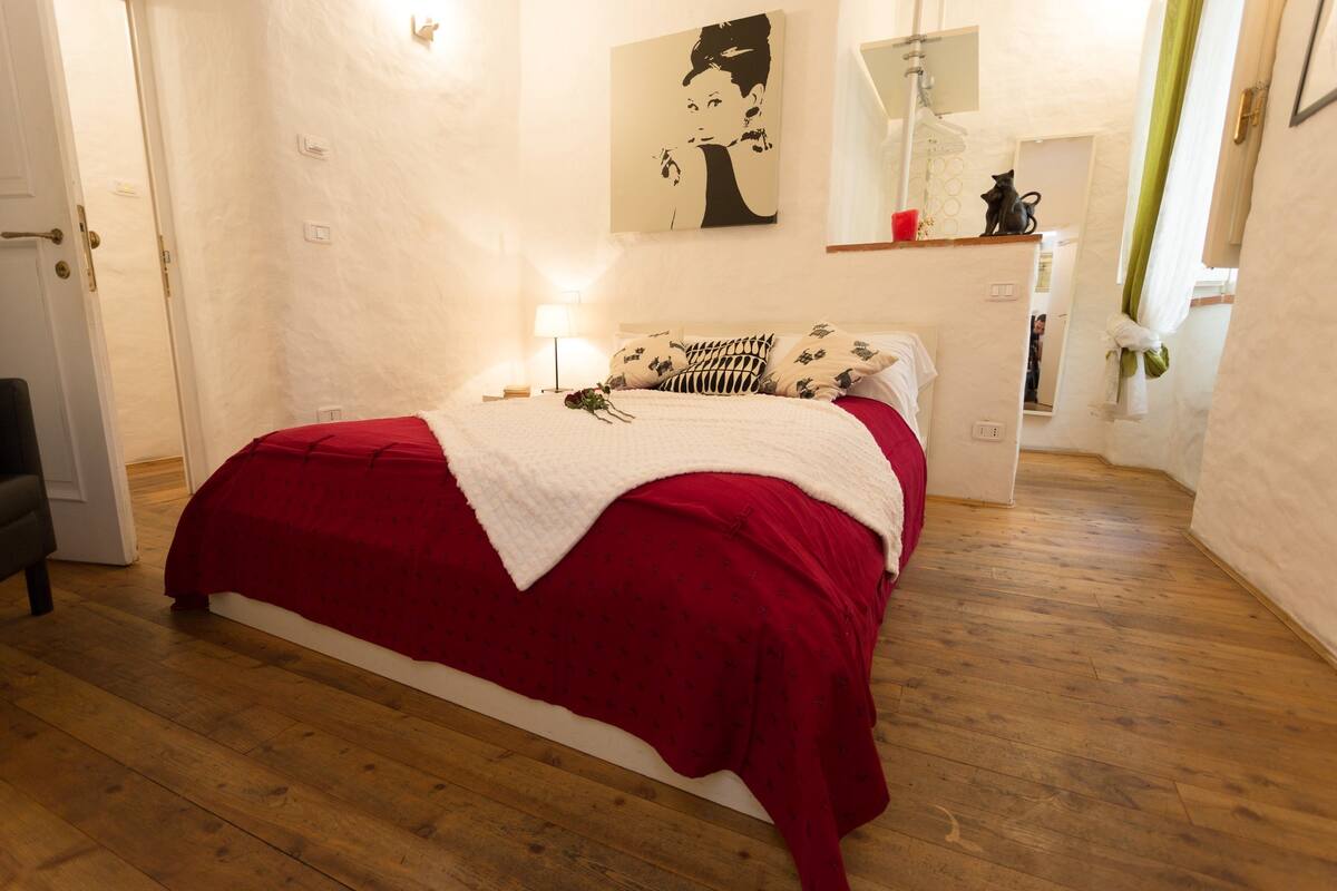 Un dormitorio con piso de madera, una cama con cubrecama rojo y blanco, una obra de arte colgada en la pared y una mesita de noche con una lámpara.