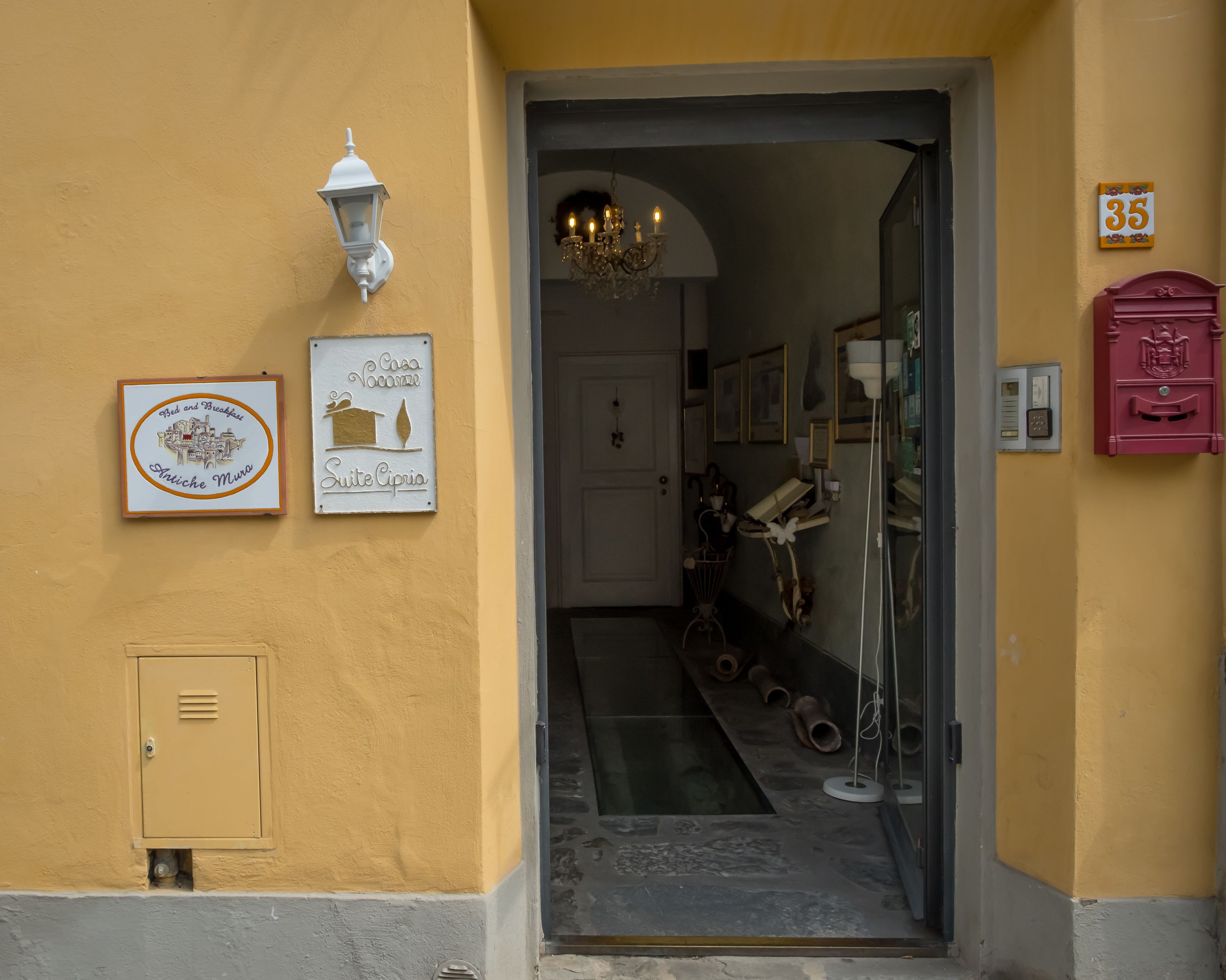 L’entrée d’un hôtel, avec un mur jaune, une enseigne indiquant « Suite Cipria », une boîte aux lettres et une porte donnant sur l’intérieur.