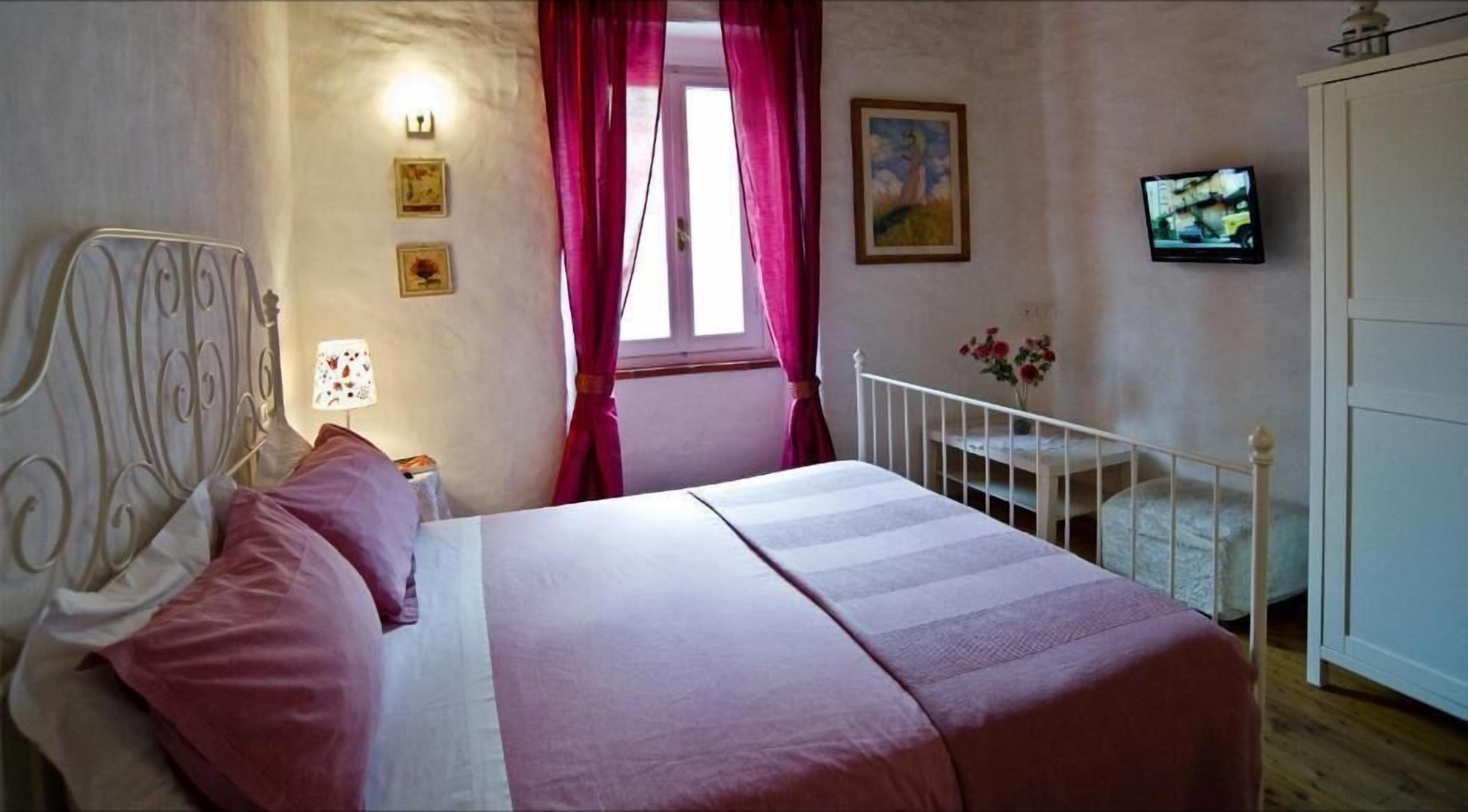 A bedroom with a bed, a nightstand, a lamp, a window with curtains, a painting, a television, and a door.
