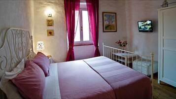 A bedroom with a bed, a nightstand, a lamp, a window with curtains, a painting, a television, and a door.