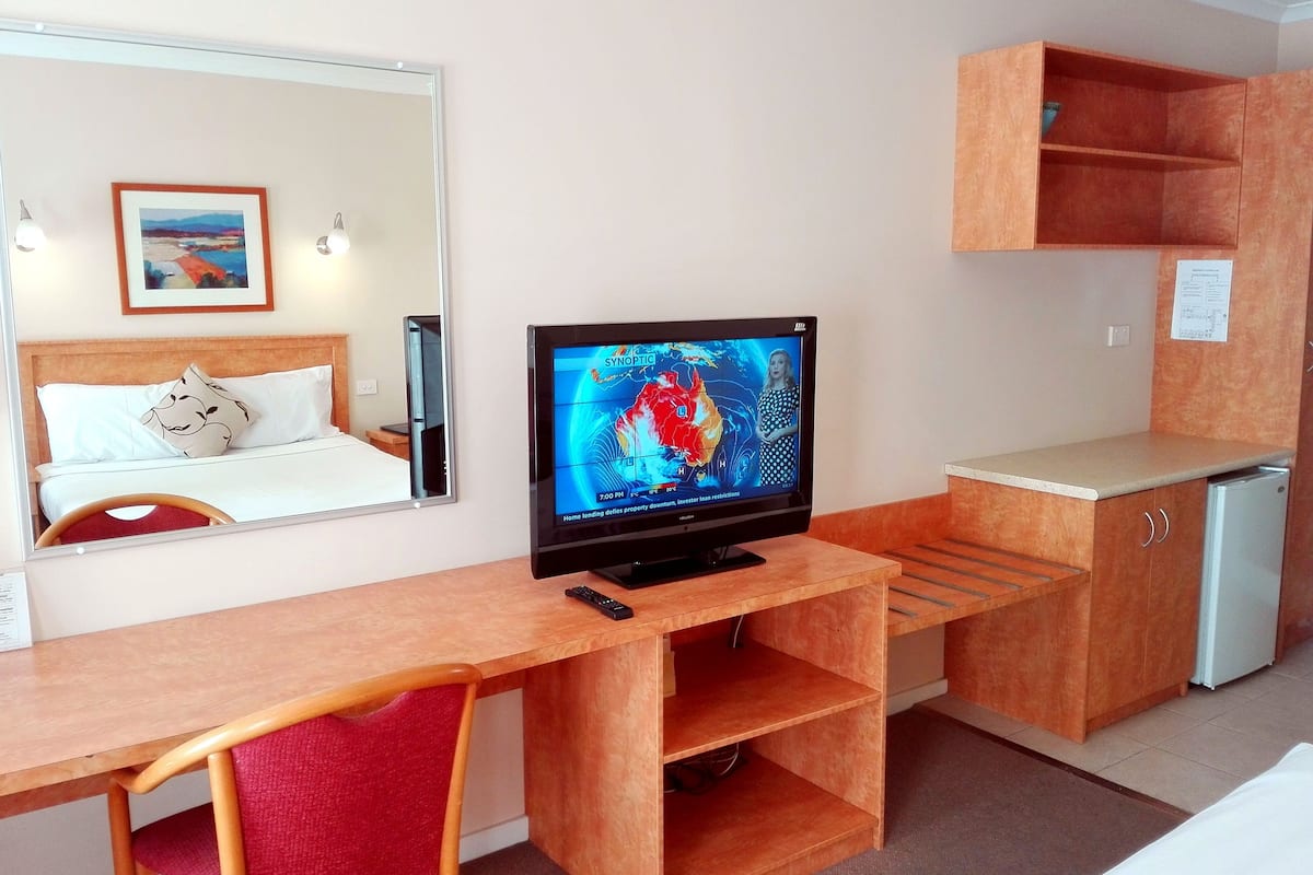 A hotel room with a wooden desk, a bed with a white headboard, a TV on the desk, a small kitchenette, and a mirror on the wall.