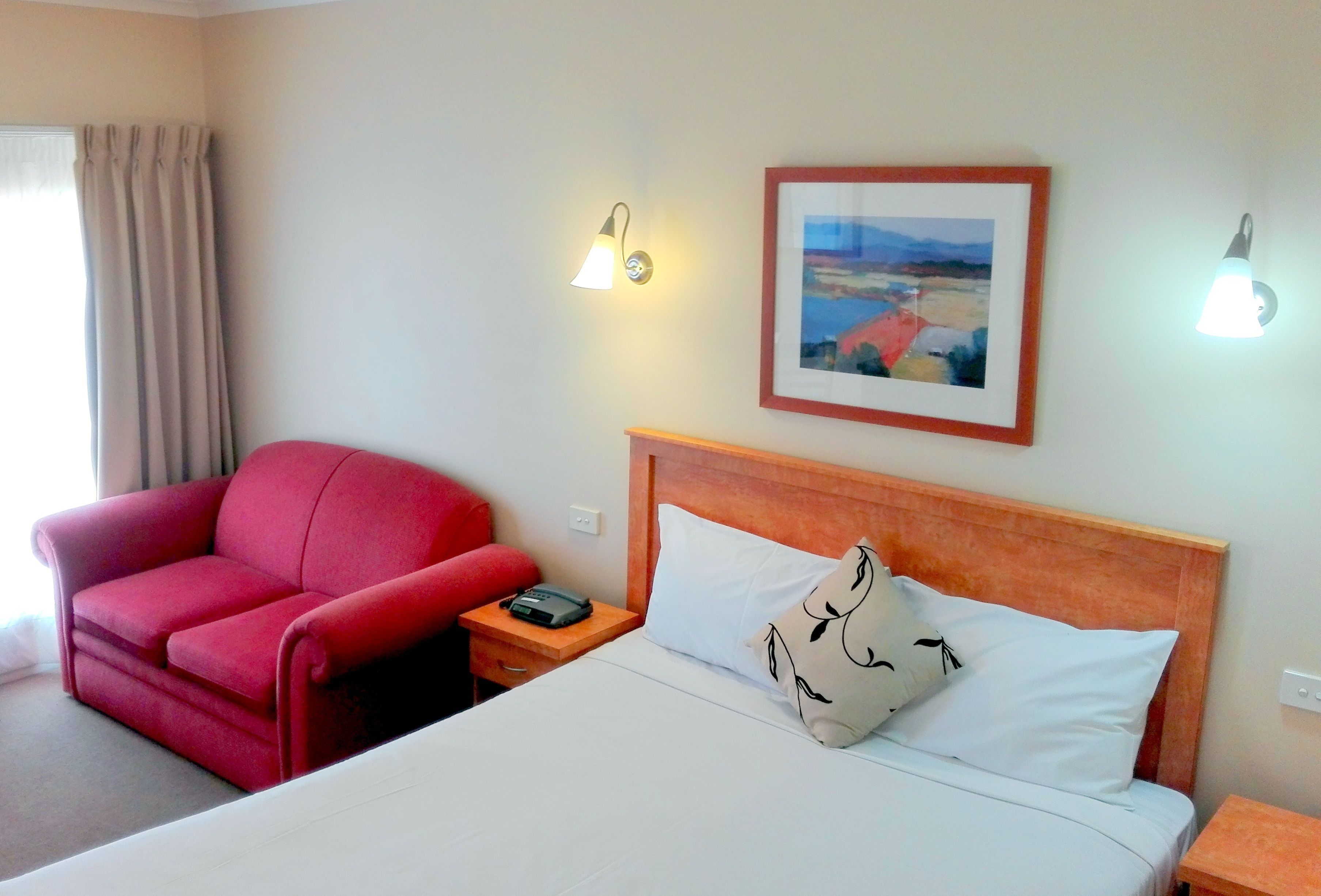A hotel room with a bed, a red armchair, a bedside table with a phone, and two wall-mounted lamps.