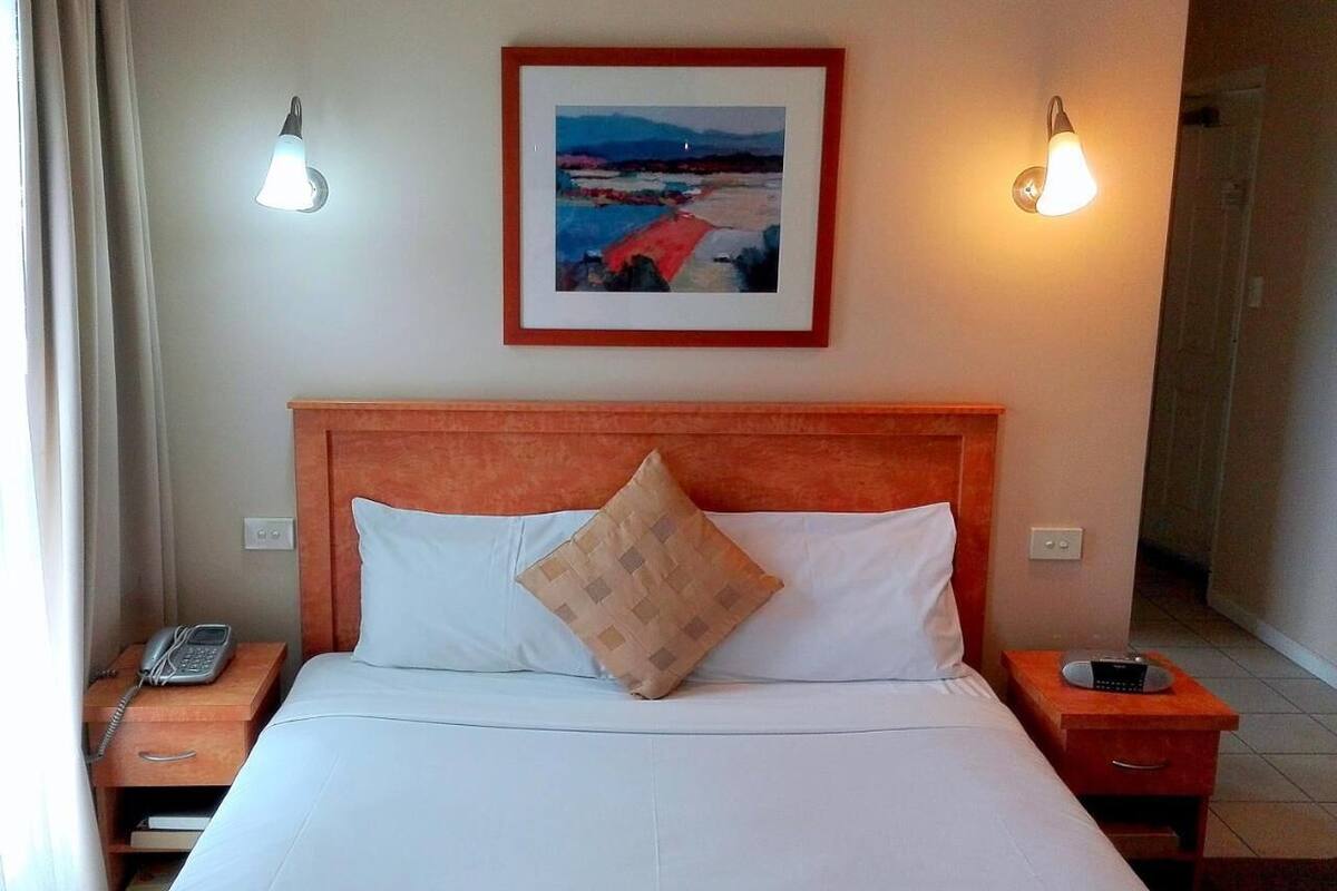 A hotel room with a bed, a nightstand with a phone, a bedside table with a radio, and a framed painting on the wall.