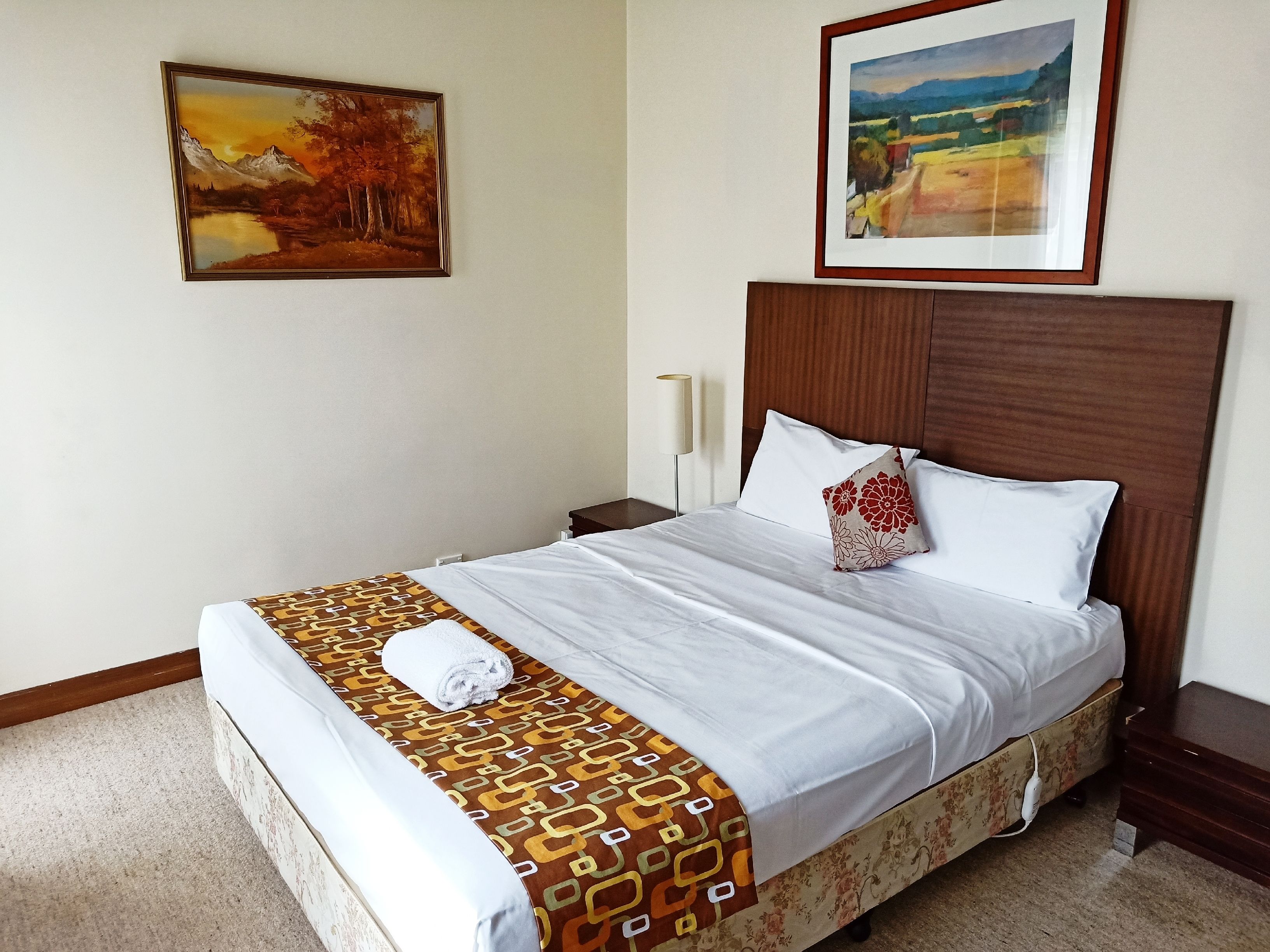 A neatly made bed with a patterned bedspread, a pillow with a floral design, and a framed landscape painting above it.