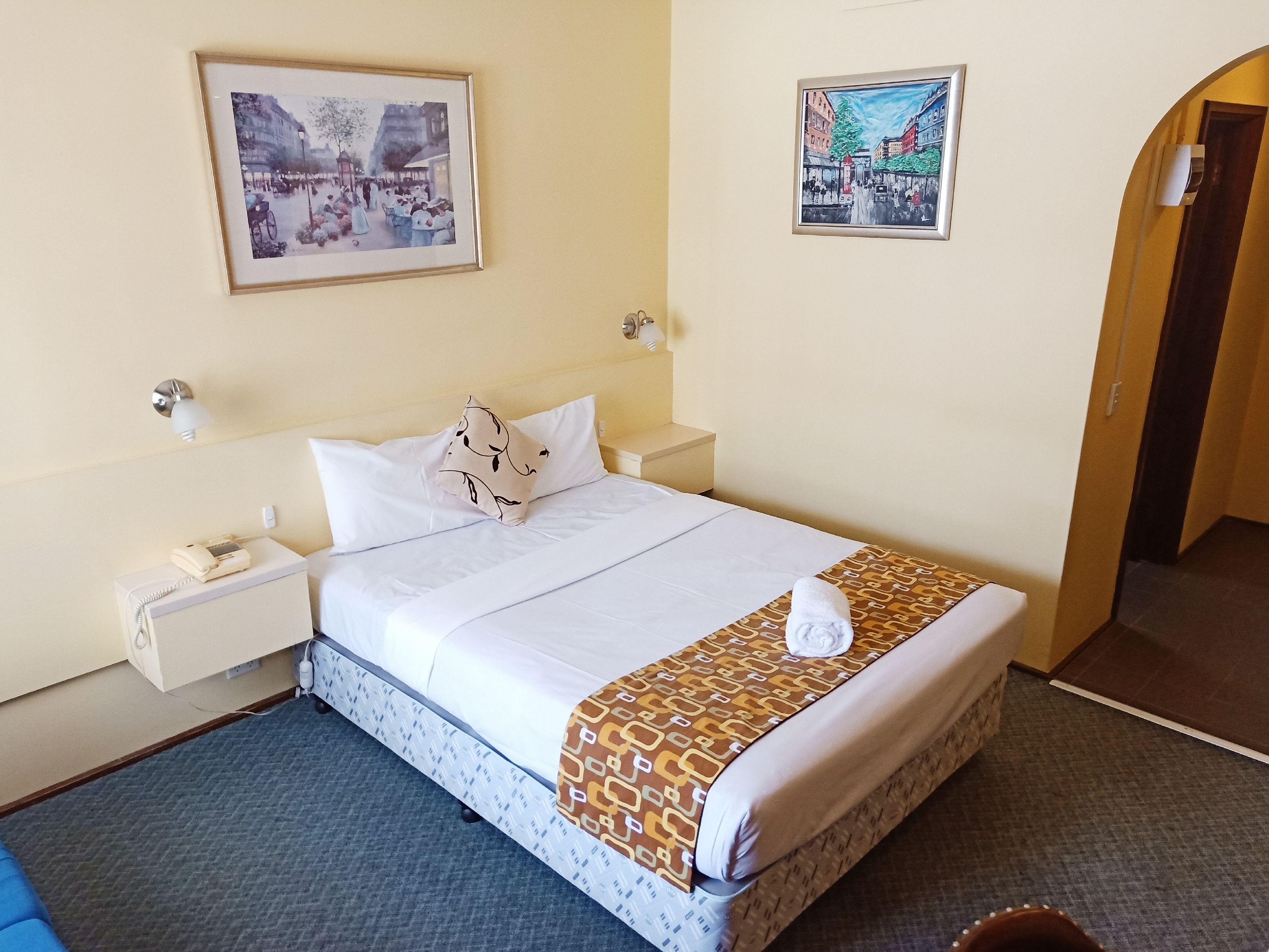 Una habitación de hotel con una cama bien tendida, un teléfono junto a la cama y dos cuadros en la pared.