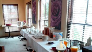 A breakfast buffet setup with various food items and beverages, a table with cups and a pitcher, and large windows with blinds. - Glen Inn (Glen Waverley)