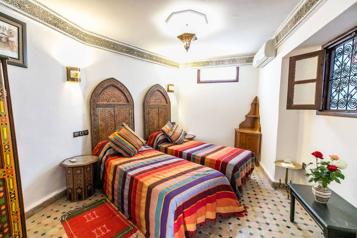 A room with two beds, patterned rugs, a small table, and decorative wall elements.