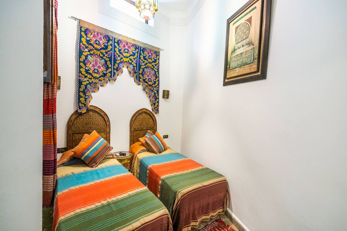 A room with two beds, colorful striped bedding, a decorative headboard, a small table, and a framed picture on the wall.