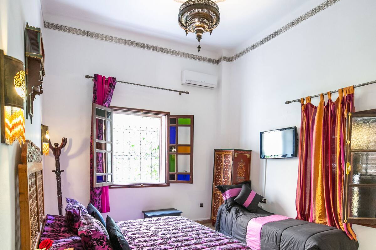 A bedroom with a bed, a television, a window with curtains, a ceiling light, and a patterned rug.