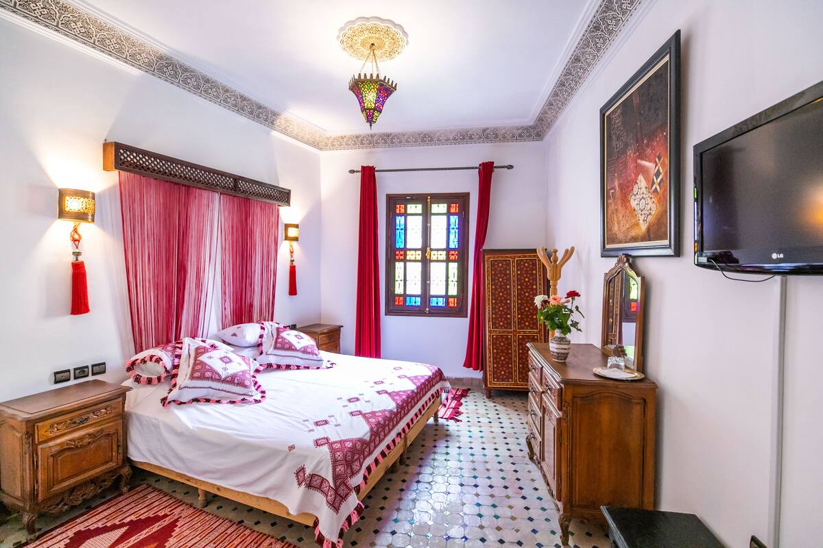 A bedroom with a bed, a wooden dresser, a TV, and a window with red curtains.
