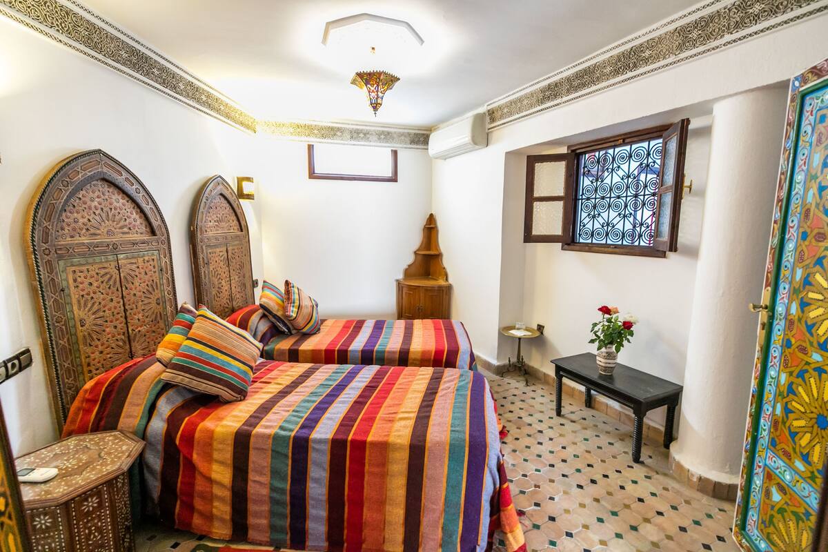 A room with two beds, a small table, a decorative lamp, and a window with intricate patterns.