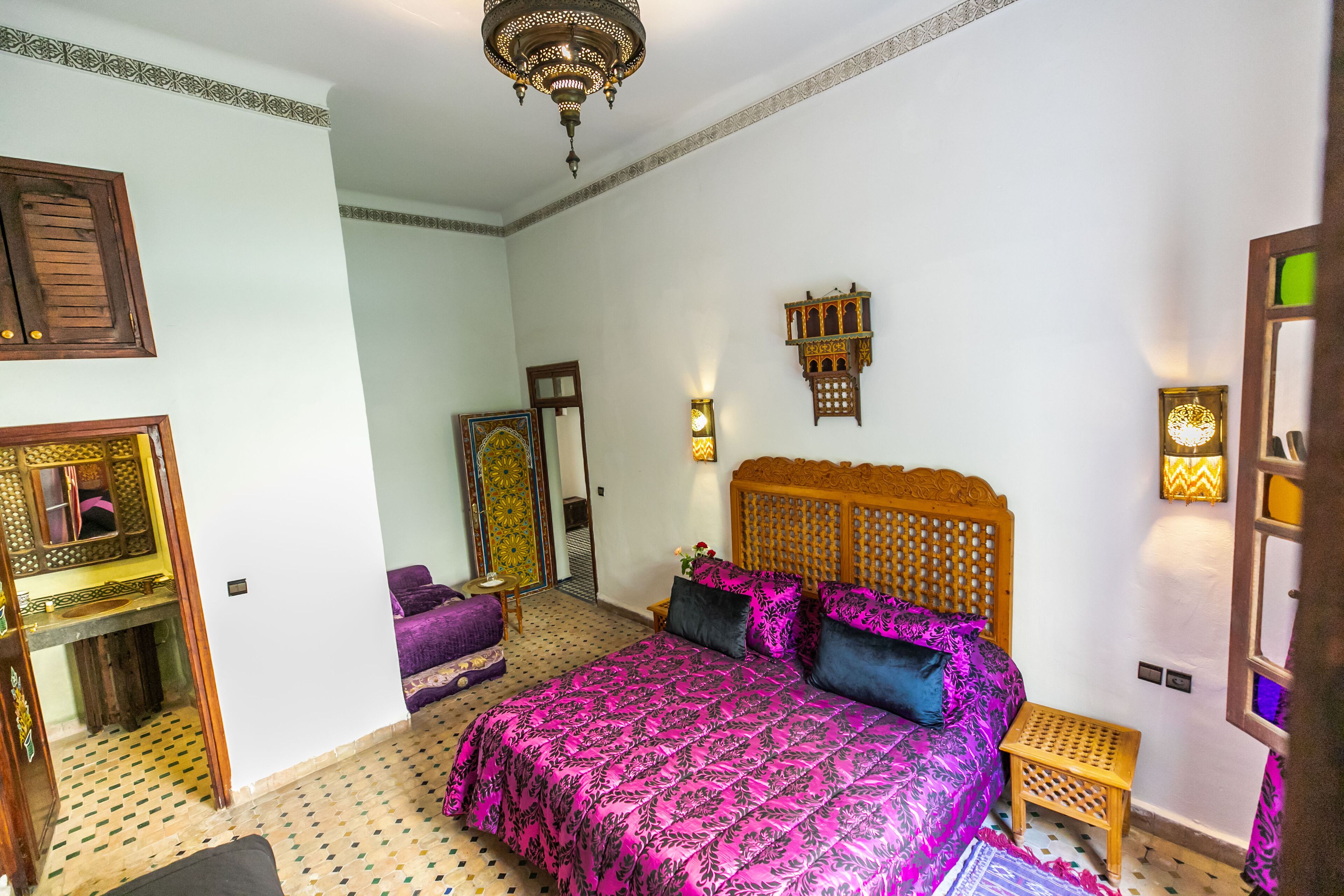 A bedroom with a large bed, a wooden headboard, a small wooden nightstand, a mirror, and a patterned floor.