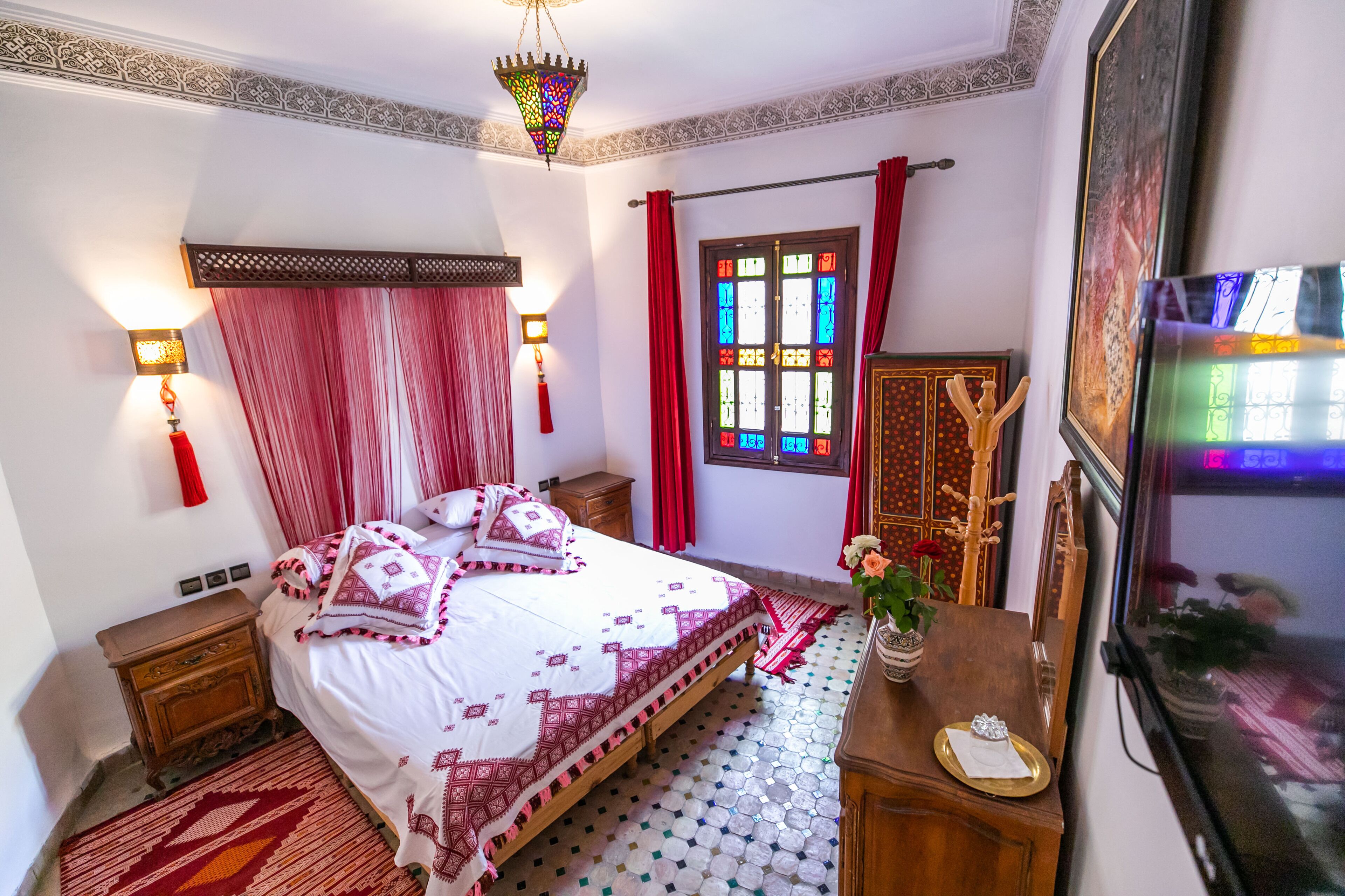 A bedroom with a bed, a nightstand, a dresser, a mirror, a window with red curtains, and a chandelier.