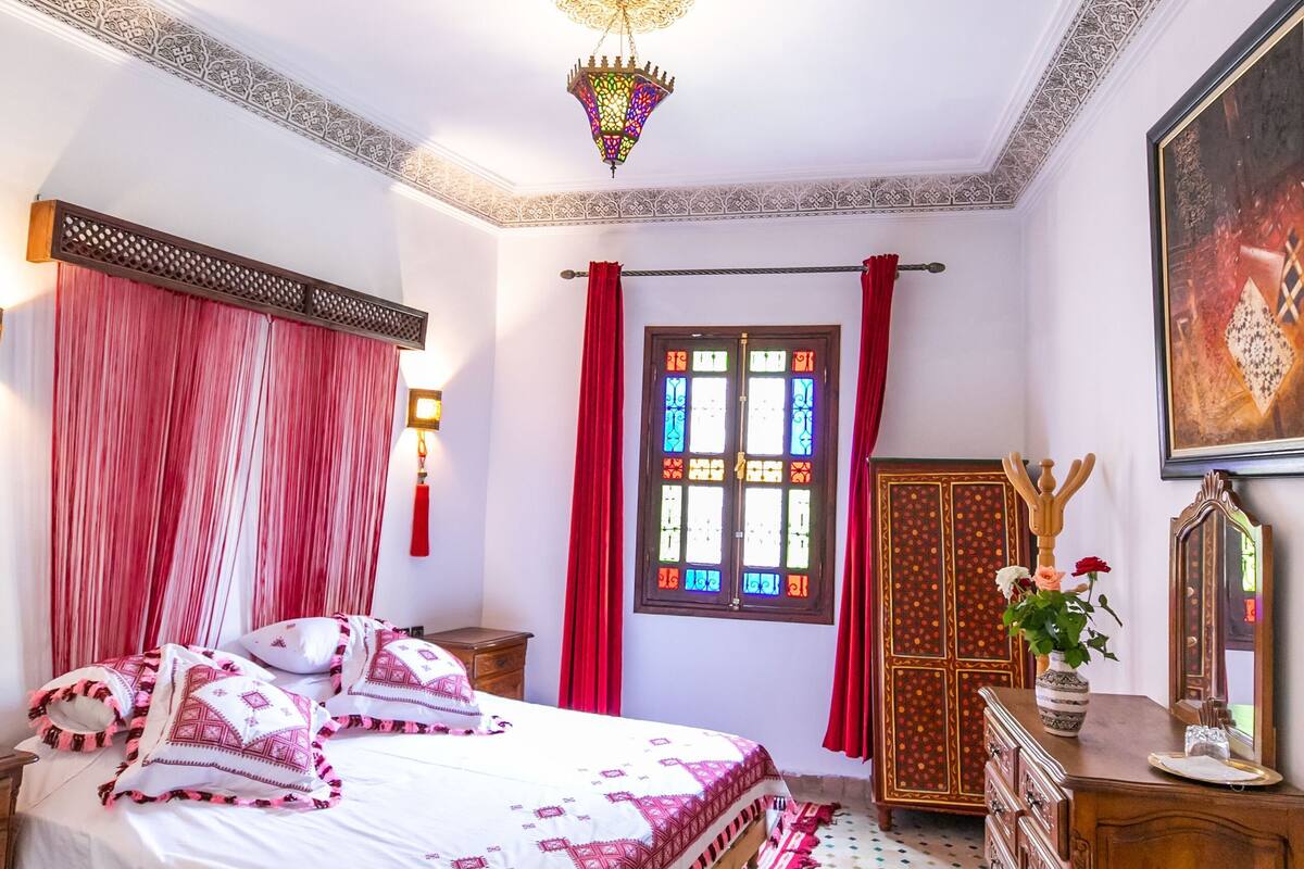 A room with a bed, a wooden cabinet, a mirror, and a window with red curtains.