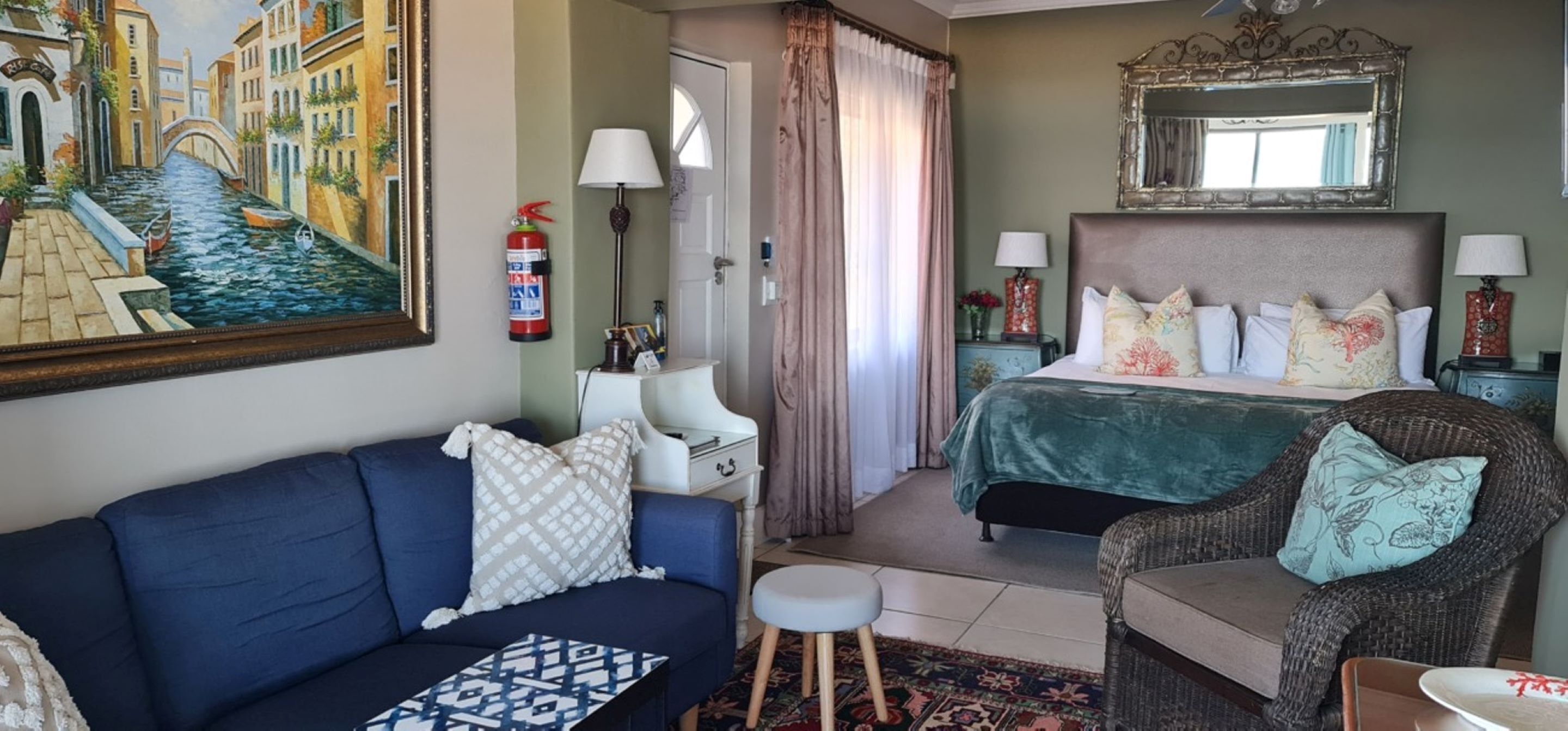 Hotelkamer met een bed, een slaapbank, een stoel, een klein krukje, een nachtkastje, een lamp, een brandblusser, een spiegel en een schilderij van een gracht.