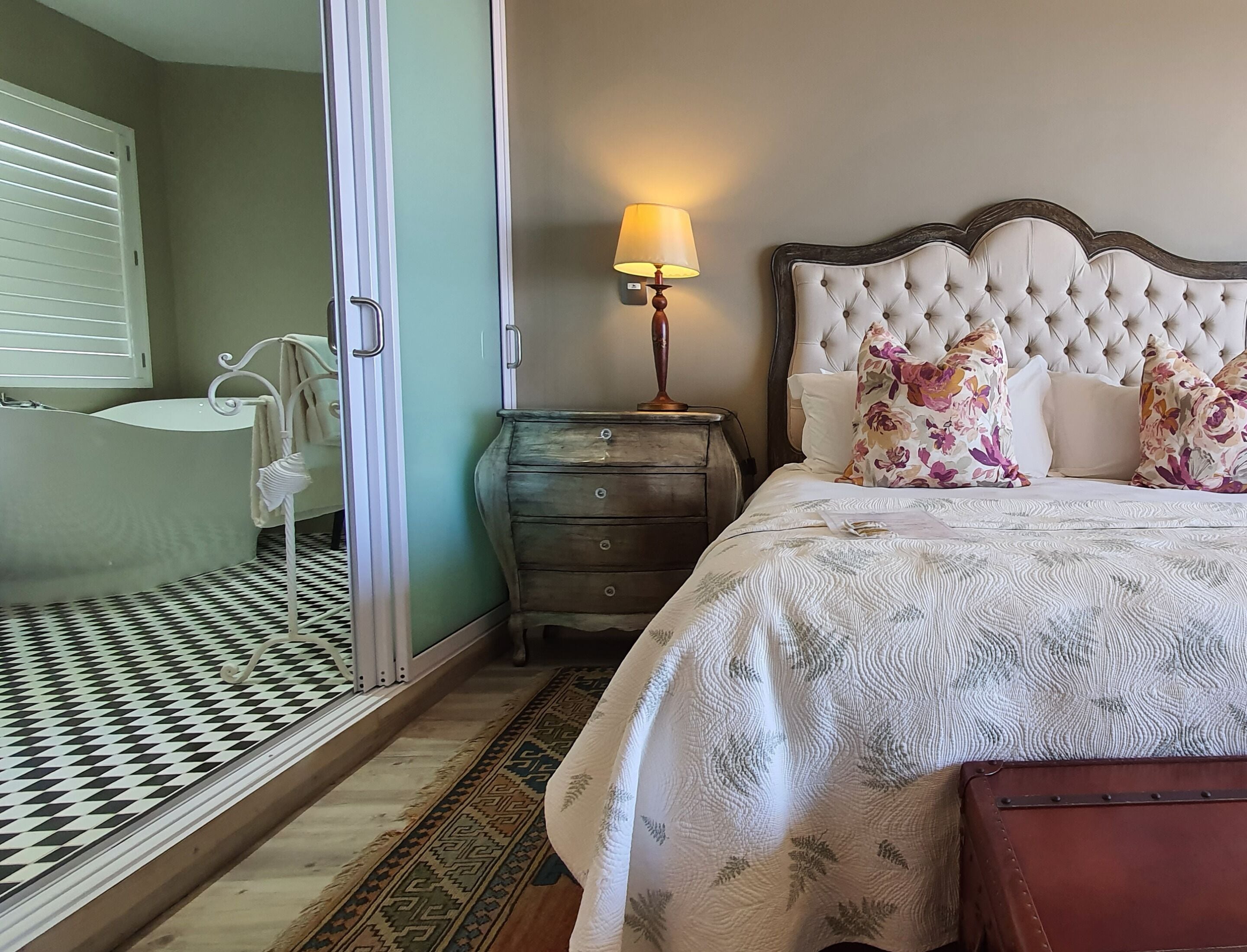 A bedroom with a bed, a nightstand, a lamp, a dresser, a bathroom with a bathtub, and a patterned floor.