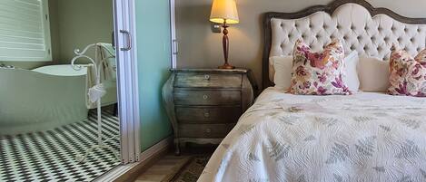 A bedroom with a bed, a nightstand, a lamp, a dresser, a bathroom with a bathtub, and a patterned floor.