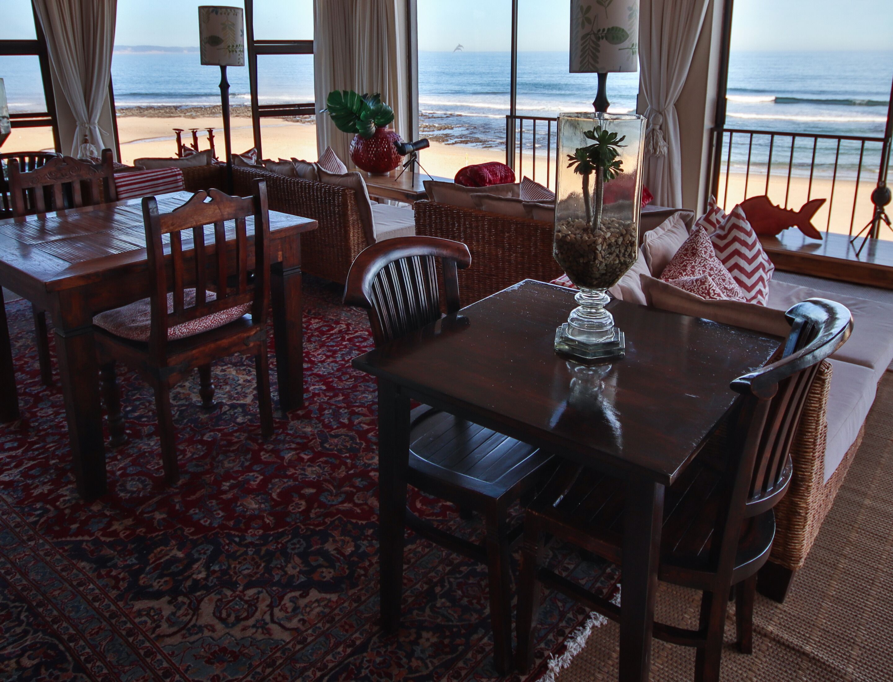 A room with a view of the beach, featuring a dining area with wooden furniture and a patterned rug.