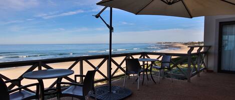 Un balcón con vista a la playa, equipado con una mesa, sillas y un paraguas grande.