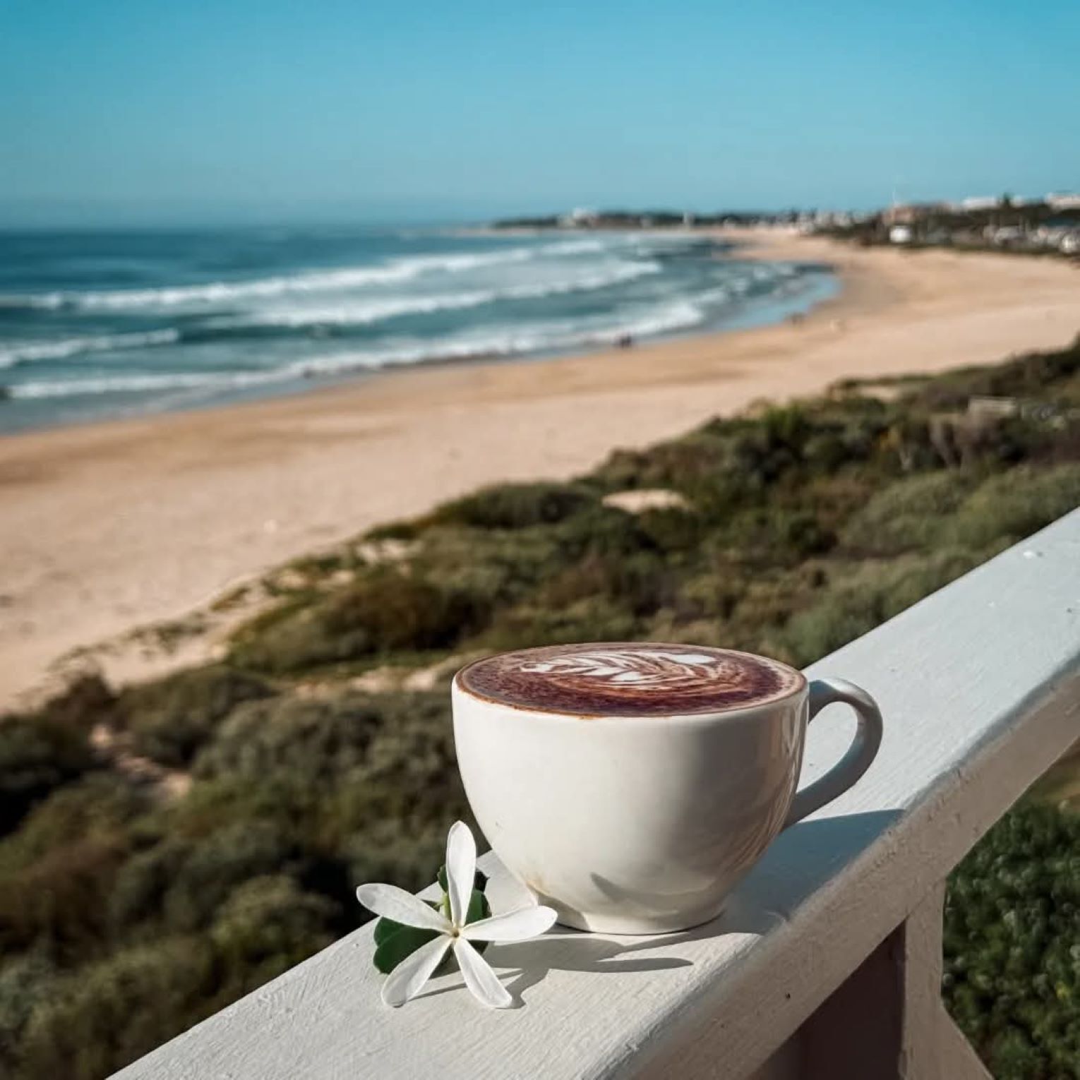A white cup of coffee with latte art on a balcony overlooking a beach.