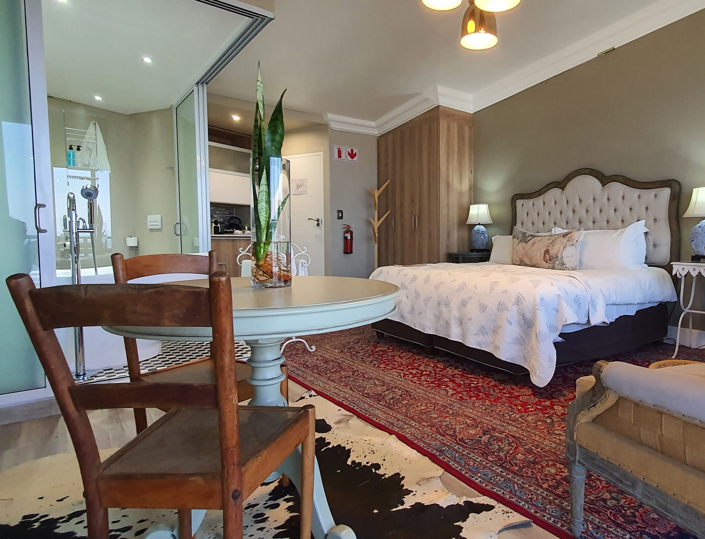 A bedroom with a bed, a round dining table, chairs, a large mirror, and a red patterned rug.