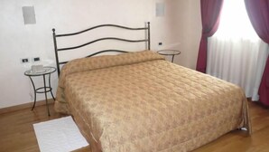Una cama individual con estructura metálica y edredón estampado en una habitación con piso de madera, una mesita redonda y un tapete blanco.