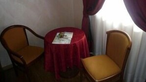 Una habitación pequeña con una mesa redonda cubierta por un mantel rojo, dos sillas de madera y una ventana con cortinas.