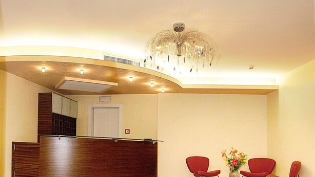 Un área de recepción con un mostrador de madera curvo, sillas rojas, una araña de cristal y un logotipo en el suelo.
