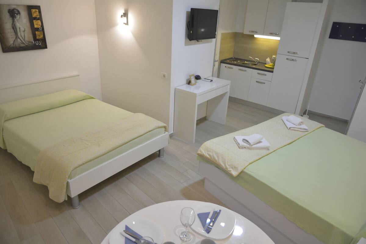 A hotel room with two single beds, a small desk with a flat-screen TV, a kitchenette, and a round dining table with a wine glass and cutlery.