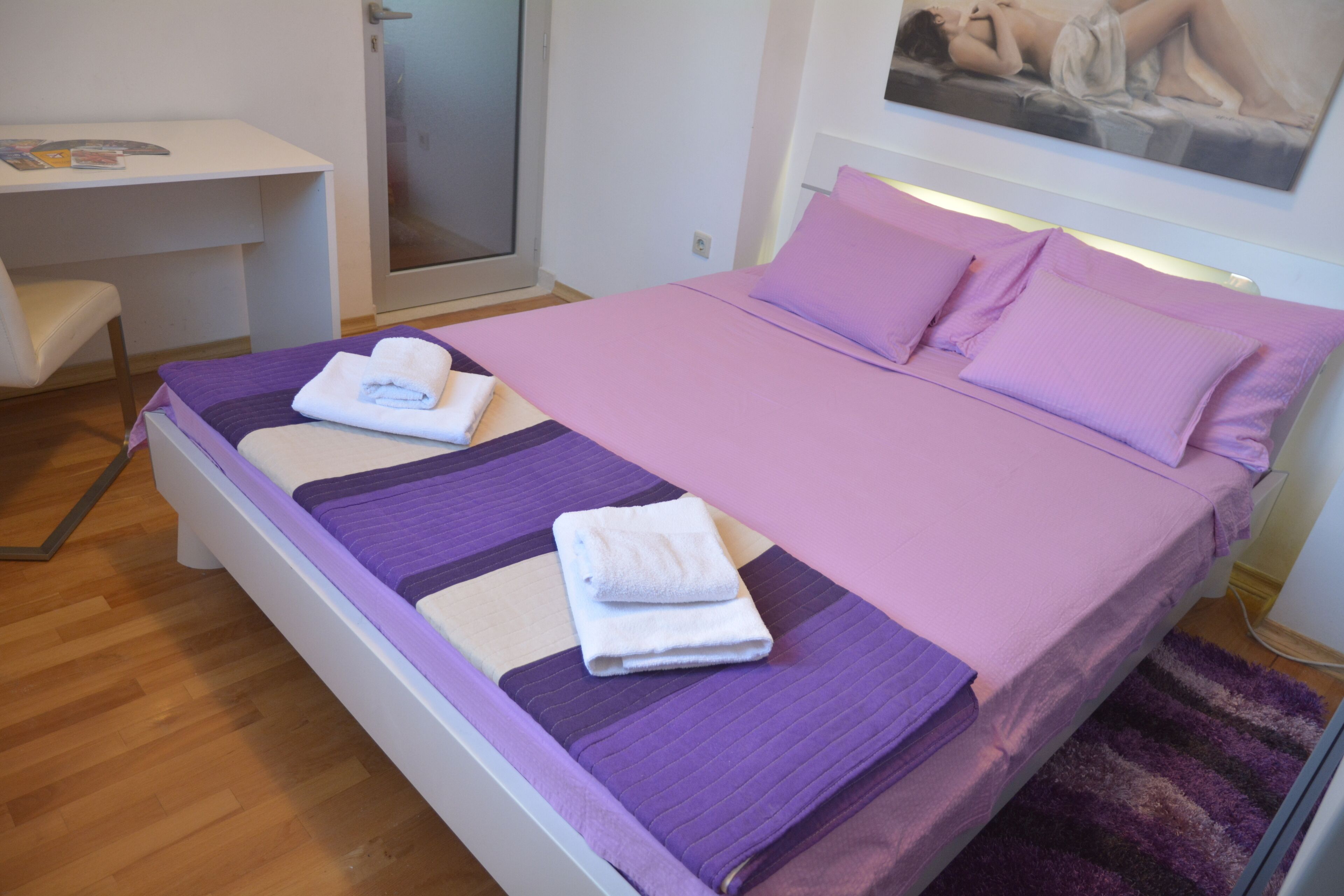 Una cama bien hecha con ropa de cama morada y toallas blancas, un escritorio pequeño con una silla, y un cuadro en la pared.