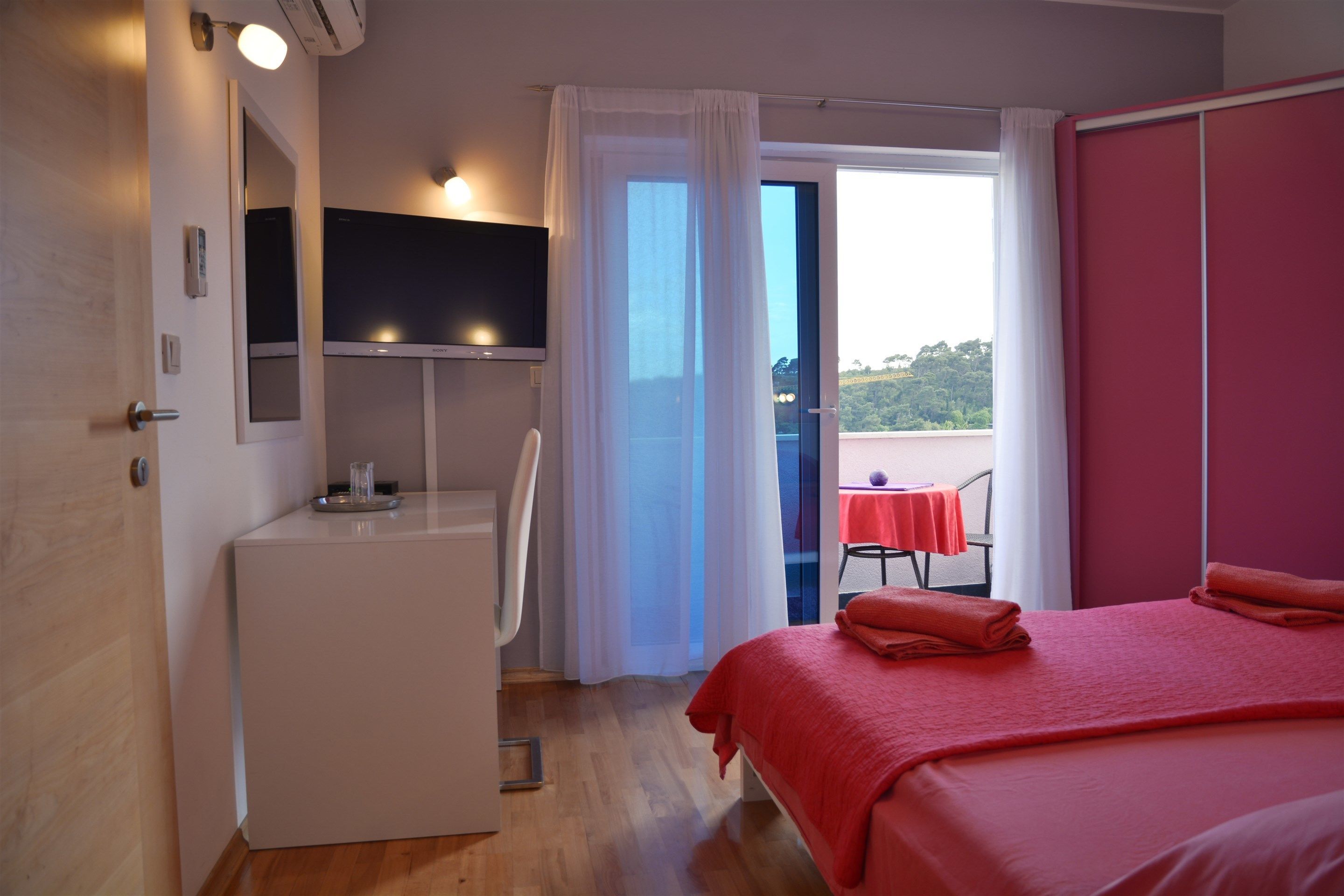 Un dormitorio con cama, escritorio, silla, televisión y balcón con mesa y sillas.