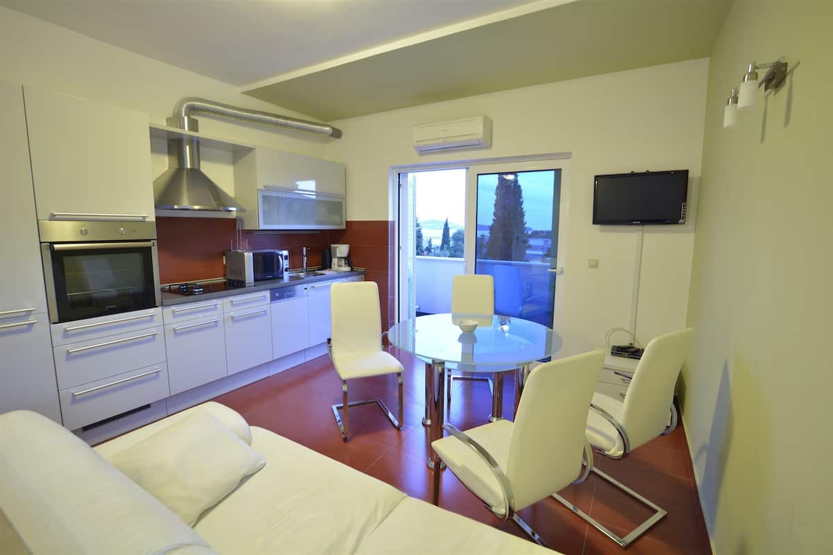A modern kitchen with white cabinets, a built-in oven, and a stainless steel range hood. There is a dining area with a glass table and four white chairs. A flat-screen TV is mounted on the wall, and an air conditioning unit is visible.