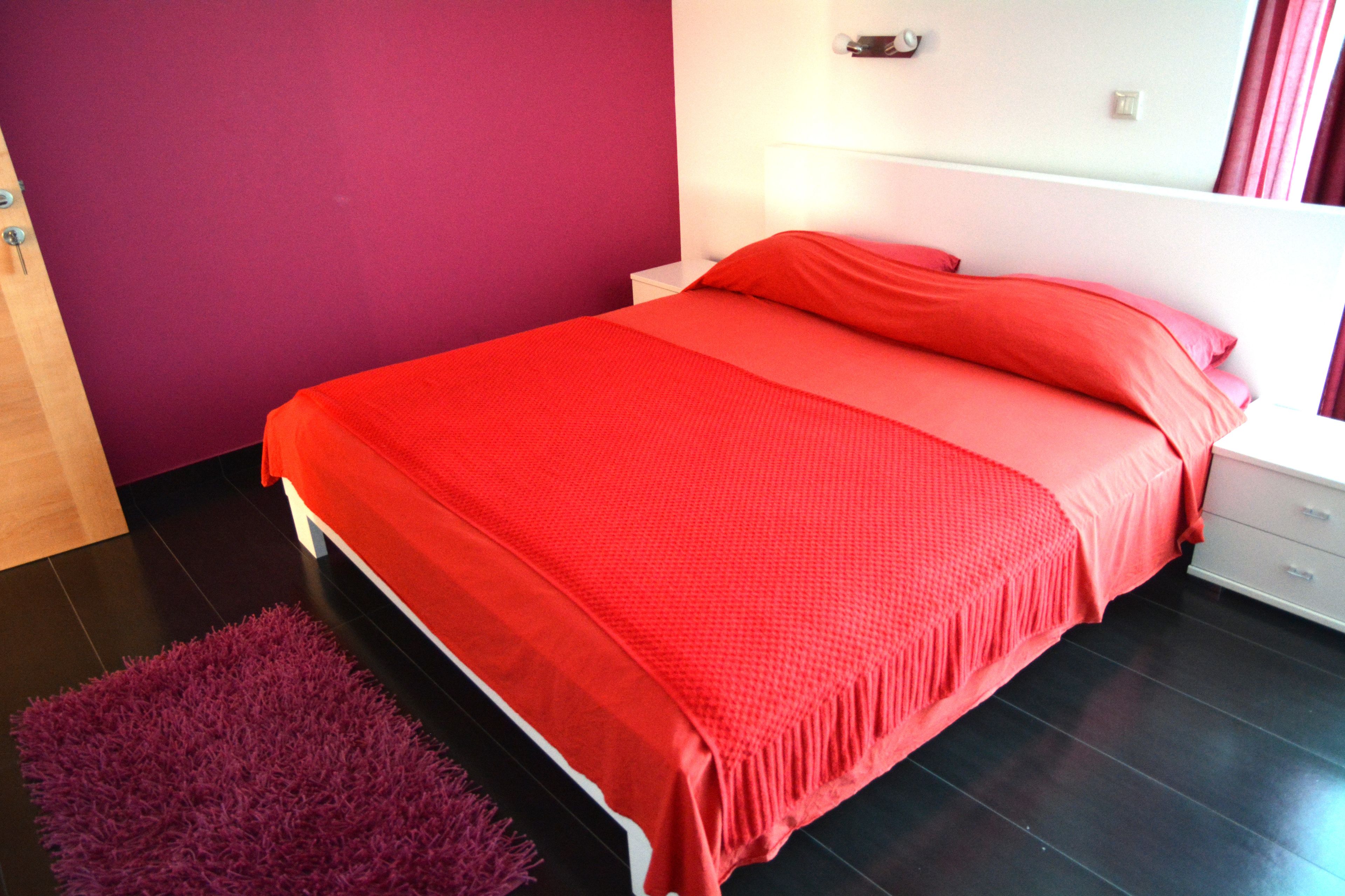 A bedroom with a bed, a nightstand, a purple rug, and a white wall.