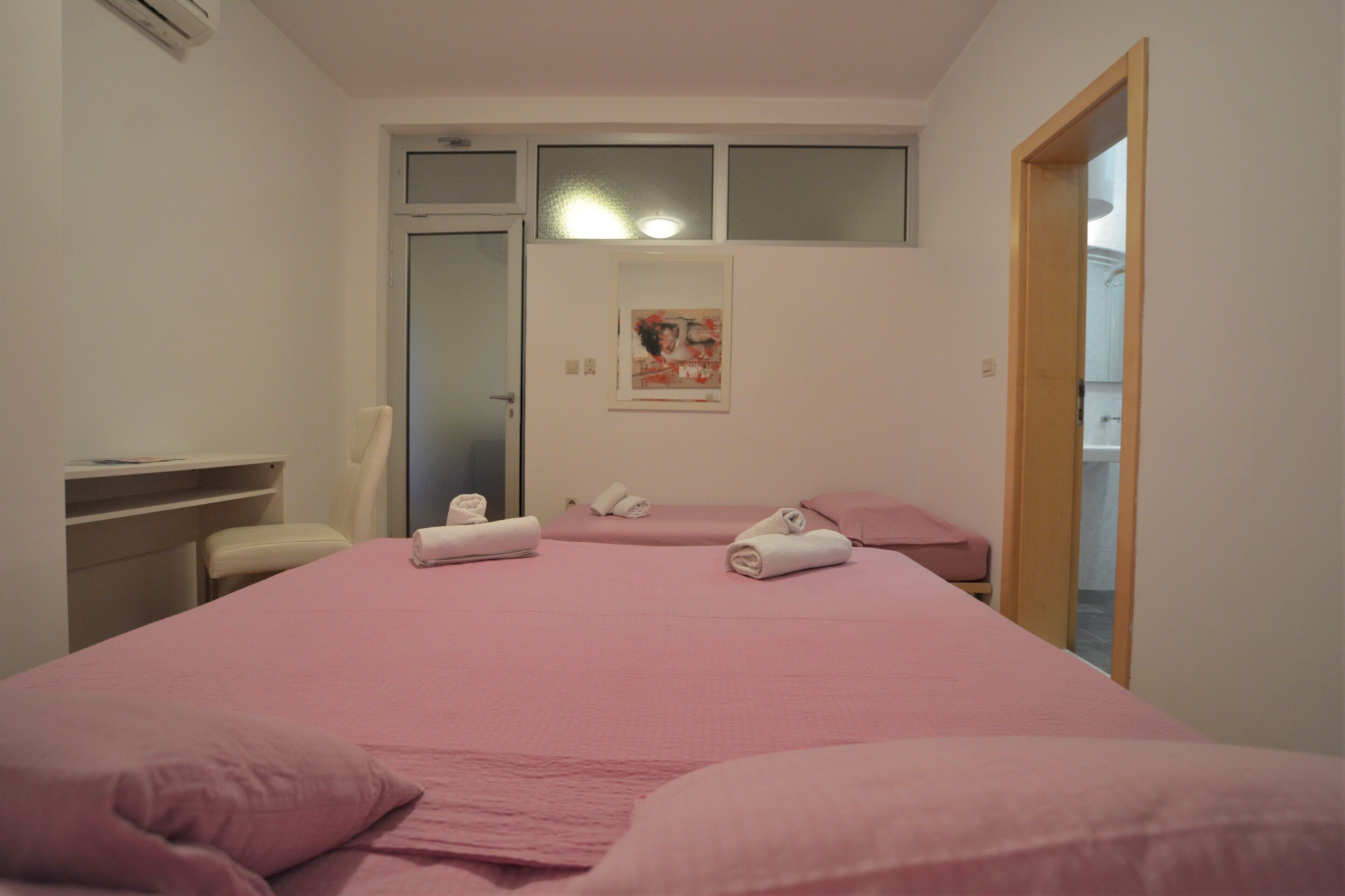 Habitación con tres camas, cada una con ropa de cama y toallas rosadas. Hay una silla y una mesita de noche con una lámpara.