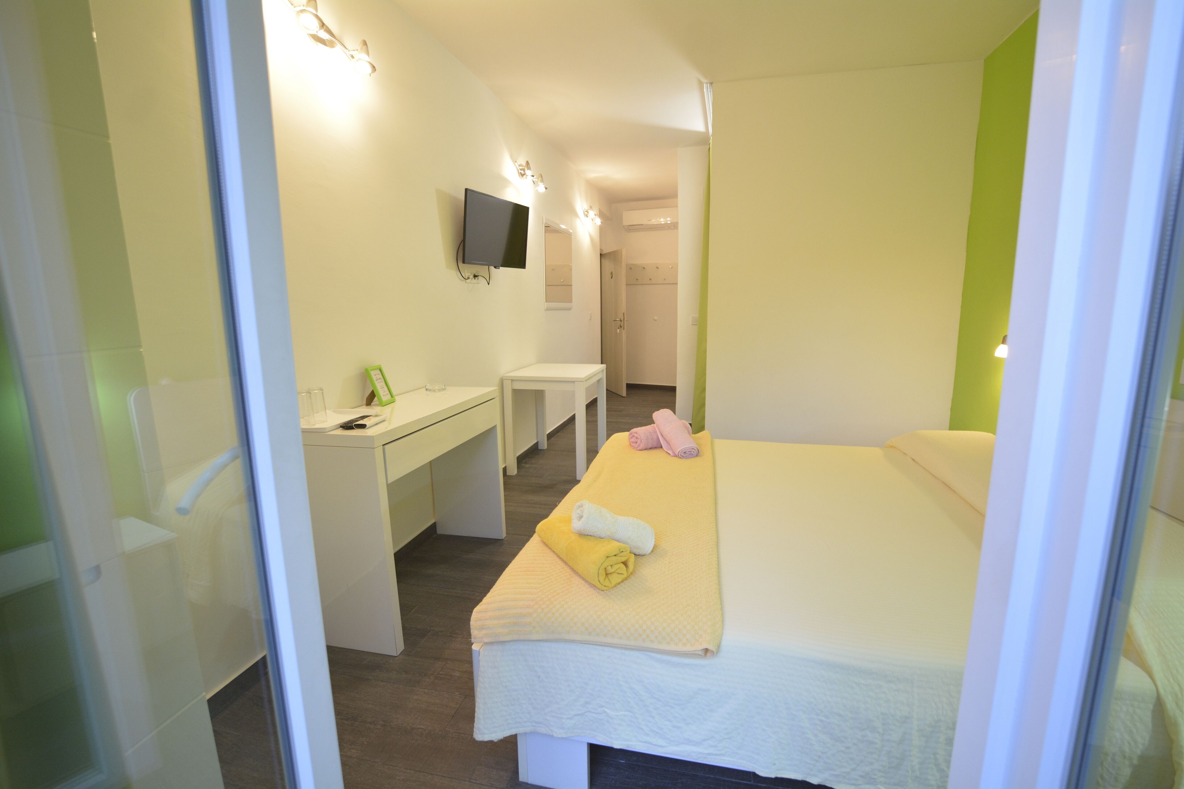A hotel room with a bed, a desk, a chair, a TV mounted on the wall, and a bathroom visible through an open door.