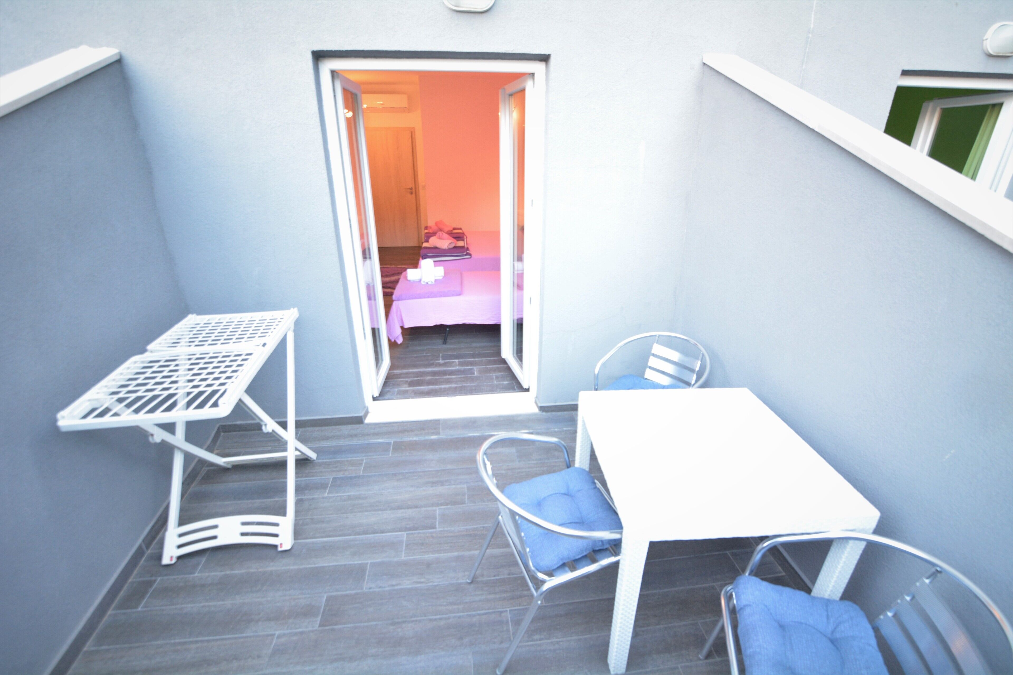 Un pequeño balcón con una mesa blanca y dos sillas, un tendedero y vista a una habitación contigua con una cama y una manta rosa.